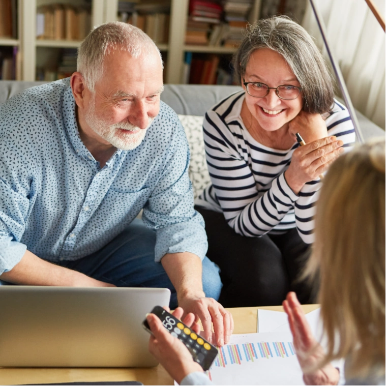 Older man and woman sitting on a couch, listening and smiling at a person who is partially visible in the foreground, with bookshelves in the background—creating a warm, welcoming atmosphere often found during real estate consultations in Glasgow.